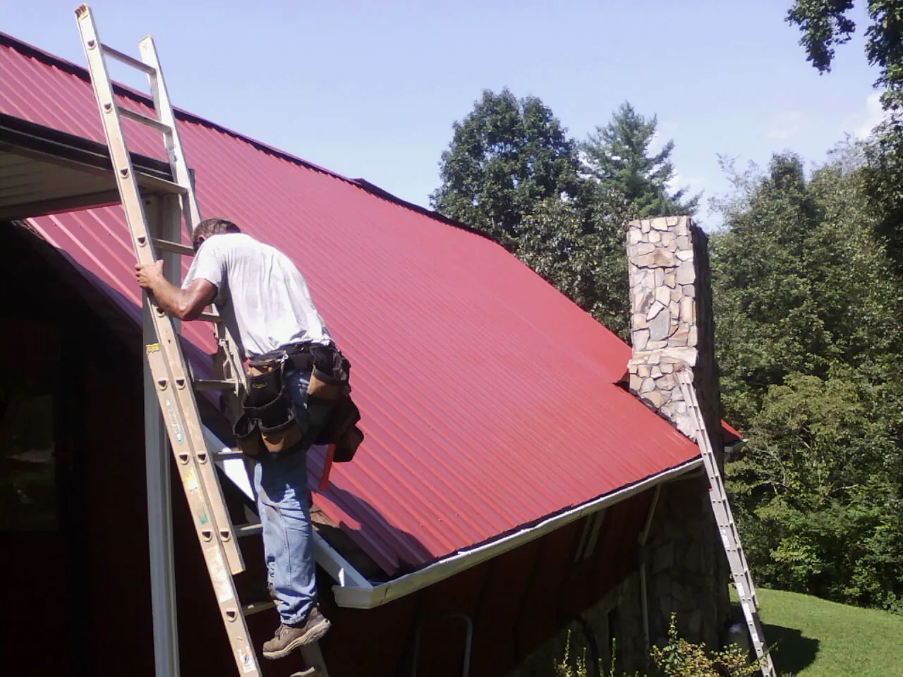 Roofer on a ladder beside a red metal roof and stone chimney — Soffit & Fascia Repair by Roofing Brothers in Griffithsville